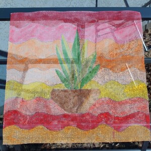 May include: A needlepoint canvas featuring a watercolor painting of a potted plant. The plant has green leaves and sits in a brown pot. The background has layers of pink, orange, yellow, and red hues, creating a sunset effect.