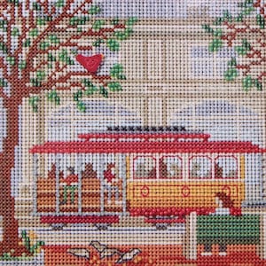 May include: Needlepoint depiction of a San Francisco cable car with a red roof and yellow body. The scene includes a tree with green leaves and a red bird, set against a building backdrop. The artwork is detailed and colorful.
