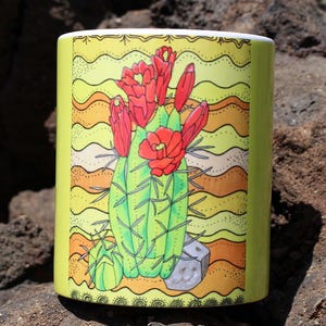 May include: A cylindrical ceramic mug featuring a hand-drawn illustration of a green cactus with red flowers. The background has yellow and orange wavy lines. The mug is set against a dark, textured surface.
