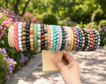 Op de afbeelding: Een verzameling van talloze armbanden met ronde edelsteenkralen, gerangschikt in een rij op een beige stoffen displaystandaard. De armbanden hebben een grote verscheidenheid aan kleuren, waaronder roze, wit, groen, blauw, zwart, geel en bruin.