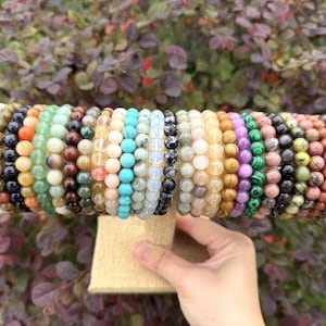May include: A collection of beaded bracelets in various colors, including red, green, blue, and purple. The bracelets are displayed on a beige stand, showcasing the diverse range of gemstones and materials used in their construction.