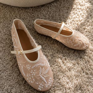 Peut inclure: Une paire de chaussures plates en maille beige avec des broderies florales blanches et de petites perles blanches. Chaque chaussure possède une fine bride crème avec une boucle en métal doré et un bout arrondi avec une semelle plate.