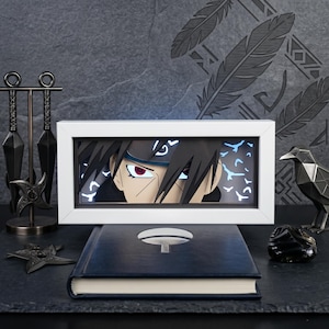 May include: A framed artwork featuring a character's face with red eyes and black hair. A dark blue book with a symbol is in front of the frame. Additional items include a metal raven sculpture, throwing stars, and kunai knives.