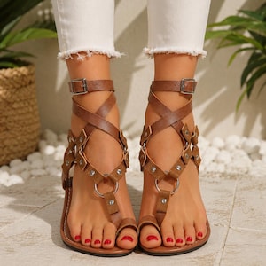 Op de afbeelding: Een paar bruine platte sandalen met een teenlus, meerdere kruisende bandjes en zilverkleurige metalen ringen en studs. De sandalen hebben een verstelbaar enkelbandje met een zilverkleurige gespsluiting en een platte, donkergekleurde zool.