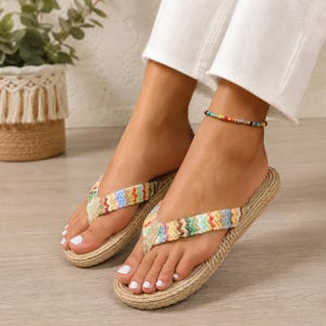 May include: A pair of flip-flops featuring a woven jute-style sole and a multi-colored chevron patterned fabric strap. The strap displays a vibrant zigzag design in shades of blue, green, yellow, orange, and pink, secured with a beige fabric toe post.