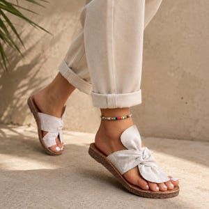 May include: A pair of light beige suede slide sandals featuring a knotted front strap design and a flat, textured brown sole. The sandals are worn with beige cuffed trousers and a thin, colorful beaded anklet with small multi-colored beads.