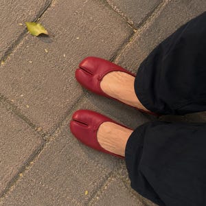 May include: A pair of deep red leather tabi-style ballet flats featuring a distinct split-toe design and a small decorative knot detail at the center of the vamp, worn with dark trousers.