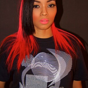 May include: A woman with long black and red hair is wearing a black t-shirt with a white geometric design.