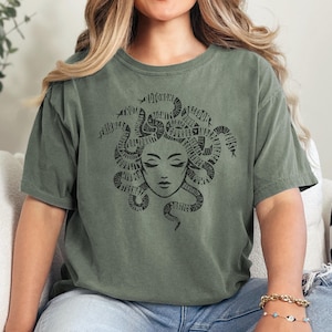 May include: Olive green t-shirt featuring a black Medusa graphic. The Medusa design includes a face with closed eyes and snakes for hair. The word "JUSTICE" is visible in the design. The shirt is made of a soft material.