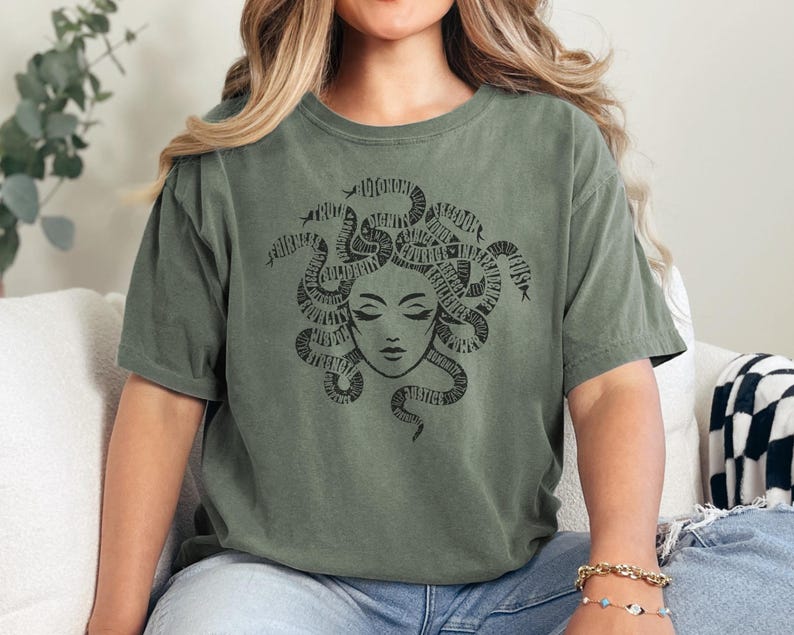 May include: Olive green t-shirt featuring a black Medusa graphic. The Medusa design includes a face with closed eyes and snakes for hair. The word "JUSTICE" is visible in the design. The shirt is made of a soft material.