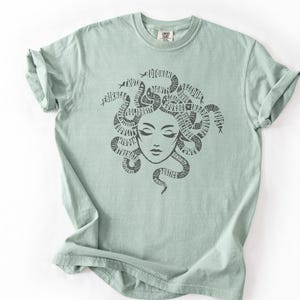 May include: A light green t-shirt featuring a Medusa graphic. The illustration depicts Medusa's face with snakes forming her hair, with words incorporated into the design. The shirt has a crew neck and short sleeves.