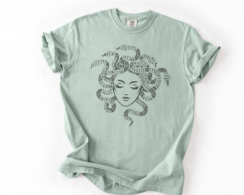 May include: A light green t-shirt featuring a Medusa graphic. The illustration depicts Medusa's face with snakes forming her hair, with words incorporated into the design. The shirt has a crew neck and short sleeves.