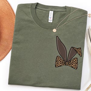 Op de afbeelding: Olijfgroen t-shirt met een afbeelding van bruine konijnenoren en een luipaardprint strikje. Een gouden ketting met een hanger en een bruine hoed liggen naast het shirt. Het shirt is gemaakt van zacht materiaal.