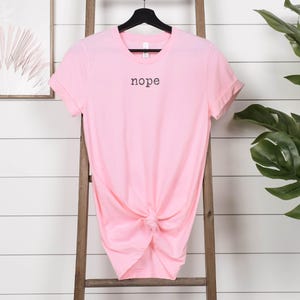 May include: A light pink t-shirt with the word "nope" printed in black. The shirt is knotted at the waist and hangs on a black hanger. The background includes a wooden ladder and a framed artwork.