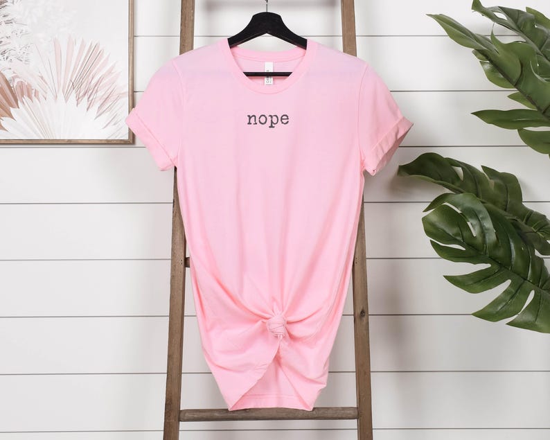 May include: A light pink t-shirt with the word "nope" printed in black. The shirt is knotted at the waist and hangs on a black hanger. The background includes a wooden ladder and a framed artwork.