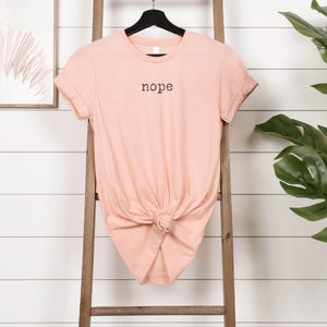 May include: A peach-colored t-shirt with the word "nope" printed in black. The short-sleeved shirt is knotted at the waist and hangs on a wooden ladder against a white-paneled wall. A black hanger supports the shirt.