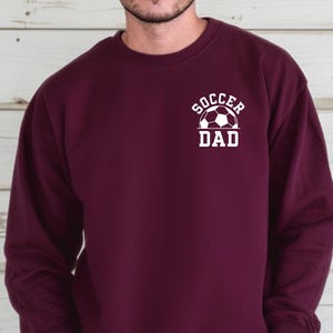 Pu&ograve; includere: Felpa bordeaux con la scritta bianca "Soccer Dad" e una grafica di pallone da calcio. La felpa girocollo a maniche lunghe &egrave; in tinta unita. Un capo casual per gli appassionati di calcio.