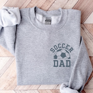 Pu&ograve; includere: Una felpa grigio melange con la scritta "SOCCER DAD" e un disegno di un pallone da calcio. Il testo e il disegno sono grigio scuro. L'indumento &egrave; piegato su una superficie di legno chiaro.