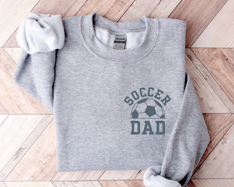 Pu&ograve; includere: Una felpa grigio melange con la scritta "SOCCER DAD" e un disegno di un pallone da calcio. Il testo e il disegno sono grigio scuro. L'indumento &egrave; piegato su una superficie di legno chiaro.