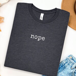 May include: Dark gray t-shirt with the word "nope" embroidered in white. The shirt is folded neatly on a white surface, with a brown hat and blue jeans visible in the background. A casual, everyday wear item.