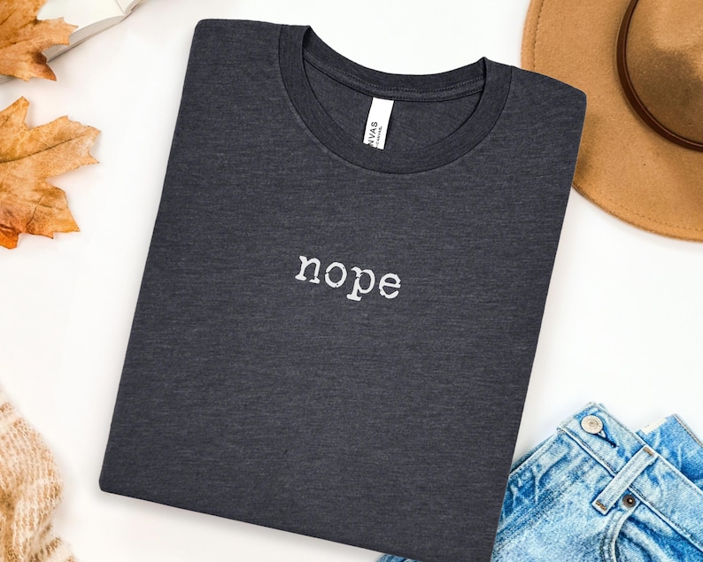 May include: Dark gray t-shirt with the word "nope" embroidered in white. The shirt is folded neatly on a white surface, with a brown hat and blue jeans visible in the background. A casual, everyday wear item.