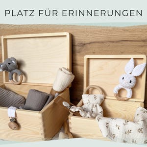 May include: Wooden memory boxes with baby items. One box has a gray elephant rattle, the other a white bunny rattle. Other items include blankets, a pacifier clip, and a small dress. The text "PLATZ F&Uuml;R ERINNERUNGEN" is at the top.