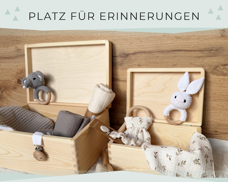 May include: Wooden memory boxes with baby items. One box has a gray elephant rattle, the other a white bunny rattle. Other items include blankets, a pacifier clip, and a small dress. The text "PLATZ F&Uuml;R ERINNERUNGEN" is at the top.