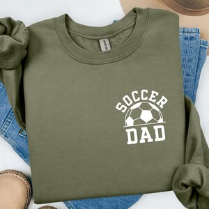 Pu&ograve; includere: Felpa verde oliva con la scritta bianca "SOCCER DAD" e una grafica di pallone da calcio. La felpa &egrave; piegata sopra jeans blu e una scarpa in pelle marrone.