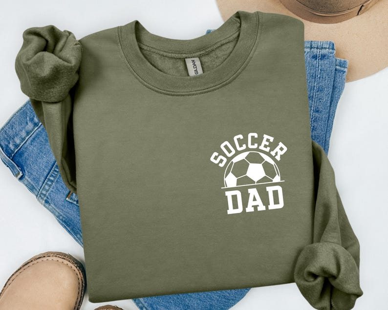 Pu&ograve; includere: Felpa verde oliva con la scritta bianca "SOCCER DAD" e una grafica di pallone da calcio. La felpa &egrave; piegata sopra jeans blu e una scarpa in pelle marrone.