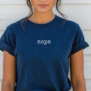 May include: A navy blue t-shirt with the word "nope" printed in white across the chest. The shirt has short sleeves and a crew neck. The model is wearing the shirt.