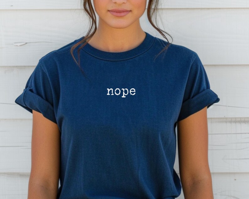 May include: A navy blue t-shirt with the word "nope" printed in white across the chest. The shirt has short sleeves and a crew neck. The model is wearing the shirt.