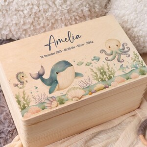 May include: A light-colored wooden box with a hinged lid, decorated with an underwater scene. The box features a whale, octopus, seahorse, and fish. The name "Amelia" is written on the lid, along with the date, time, and measurements.