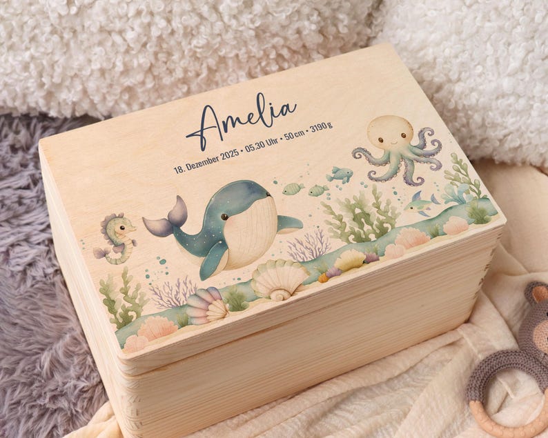 May include: A light-colored wooden box with a hinged lid, decorated with an underwater scene. The box features a whale, octopus, seahorse, and fish. The name "Amelia" is written on the lid, along with the date, time, and measurements.