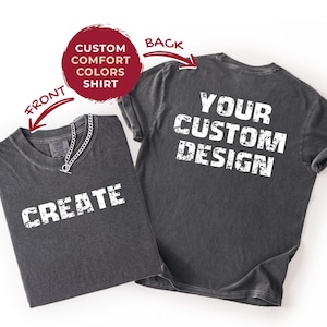 Può includere: Due magliette grigio antracite. La maglietta anteriore ha la parola "CREATE" in lettere bianche. Una collana a catena d'argento è drappeggiata sulla maglietta. La maglietta posteriore ha le parole "YOUR CUSTOM DESIGN" in lettere bianche. Un cerchio rosso indica "CUSTOM COMFORT COLORS SHIRT".