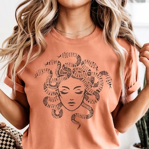 May include: A peach-colored t-shirt featuring a graphic of Medusa with snakes forming her hair. The snakes are filled with words like "truth", "freedom", and "justice". The Medusa illustration has closed eyes and a serene expression.