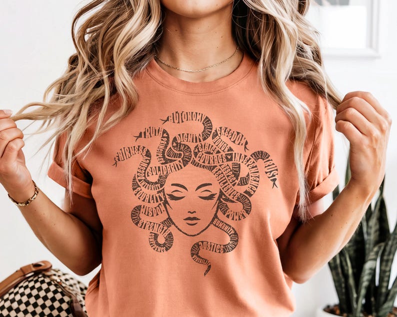 May include: A peach-colored t-shirt featuring a graphic of Medusa with snakes forming her hair. The snakes are filled with words like "truth", "freedom", and "justice". The Medusa illustration has closed eyes and a serene expression.