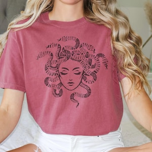 May include: A maroon t-shirt featuring a black graphic of Medusa's face, with snakes forming her hair. The word "HONOR" is visible in the design. The shirt has short sleeves and a crew neck.
