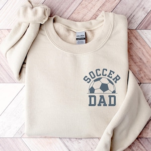 Pu&ograve; includere: Una felpa color crema con la scritta "SOCCER DAD" stampata in grigio, insieme a una grafica di pallone da calcio. La felpa &egrave; realizzata in un materiale morbido e ha uno scollo tondo.