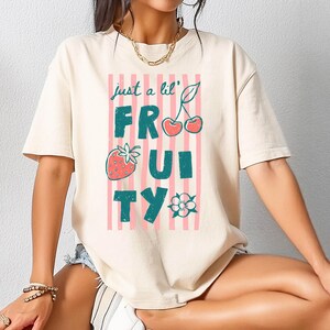 Könnte beinhalten: Ein cremefarbenes Kurzarm-T-Shirt mit einem Grafikdruck aus vertikalen rosa Streifen und dem Text just a lil' FRUITY in dunklem Petrol. Das Design enthält eine rote Erdbeere, ein Paar rote Kirschen und eine kleine weiße Blumenillustration.