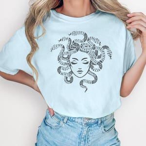 May include: Light blue t-shirt featuring a Medusa graphic. The black graphic depicts Medusa's face with snakes forming her hair, with words incorporated into the design. The shirt is paired with blue denim jeans.