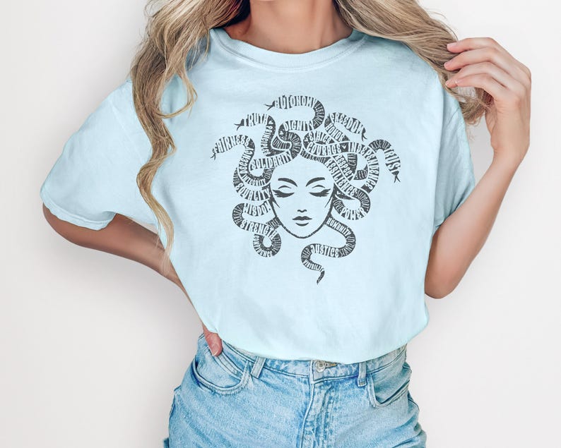 May include: Light blue t-shirt featuring a Medusa graphic. The black graphic depicts Medusa's face with snakes forming her hair, with words incorporated into the design. The shirt is paired with blue denim jeans.