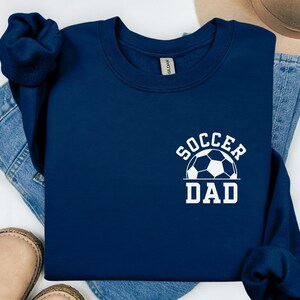 Pu&ograve; includere: Felpa blu navy con la scritta bianca "SOCCER DAD" e una grafica di pallone da calcio. La felpa &egrave; ripiegata sopra jeans blu, con un paio di scarpe beige visibili.