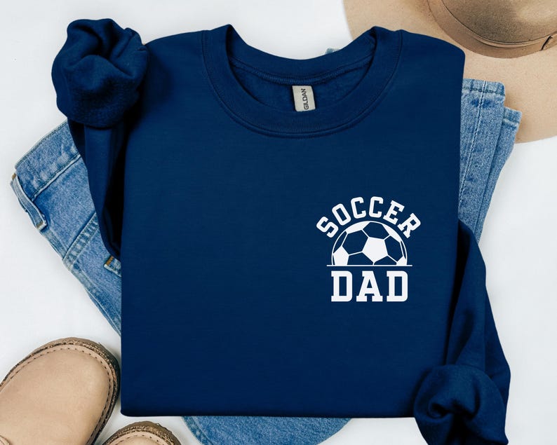Pu&ograve; includere: Felpa blu navy con la scritta bianca "SOCCER DAD" e una grafica di pallone da calcio. La felpa &egrave; ripiegata sopra jeans blu, con un paio di scarpe beige visibili.