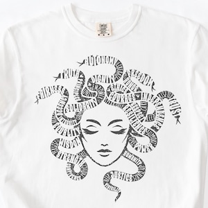 May include: White t-shirt featuring a black graphic of Medusa with words like "truth", "justice", and "courage" forming her hair. The shirt has a crew neck and short sleeves. A small tag is visible near the neckline.