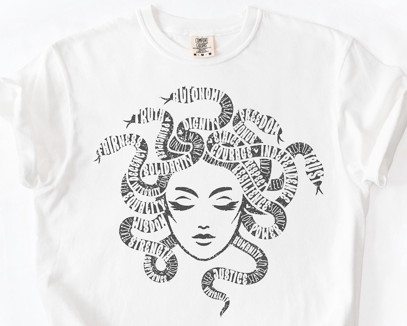 May include: White t-shirt featuring a black graphic of Medusa with words like "truth", "justice", and "courage" forming her hair. The shirt has a crew neck and short sleeves. A small tag is visible near the neckline.