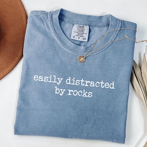 May include: A blue short-sleeved t-shirt with a crew neckline featuring the white text "easily distracted by rocks" printed on the front. The shirt has a Comfort Colors brand label at the inner neck and is made of 100% cotton fabric.