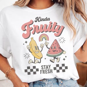 Könnte beinhalten: Weißes T-Shirt mit einem Retro-Design mit einer Banane und einer Wassermelone mit Armen und Beinen. Die Texte „Kinda Fruity“ und „Stay Fresh“ sind in einer Retro-Schriftart gehalten. Ein Regenbogen und ein Schachbrettmuster vervollständigen das Design.
