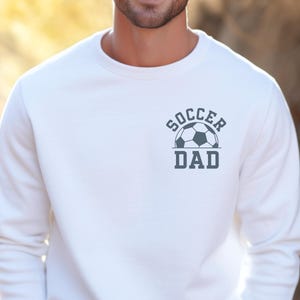 Pu&ograve; includere: Felpa bianca con la scritta "SOCCER DAD" e una grafica di pallone da calcio grigio. La felpa &egrave; a girocollo e sembra essere realizzata in un materiale morbido. Il disegno &egrave; centrato sul petto.