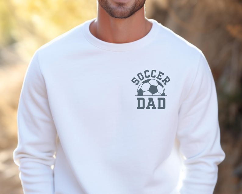 Pu&ograve; includere: Felpa bianca con la scritta "SOCCER DAD" e una grafica di pallone da calcio grigio. La felpa &egrave; a girocollo e sembra essere realizzata in un materiale morbido. Il disegno &egrave; centrato sul petto.