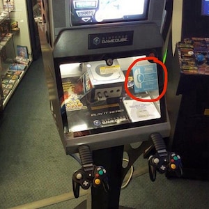 May include: A grey Nintendo GameCube retail demo kiosk featuring a top screen displaying Star Fox, a central console unit, and two black controllers hanging from the front. A blue informational card with a phone icon is visible inside the display case.
